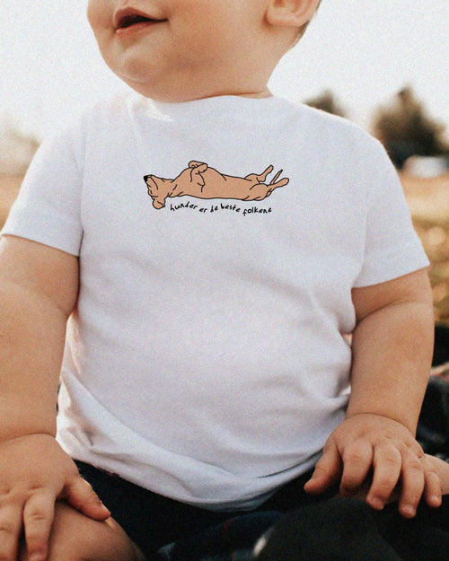 Baby med hvit T-skjorte med motiv av hund og teksten "hunder er de beste folkene", sittende ute i sola og smile.